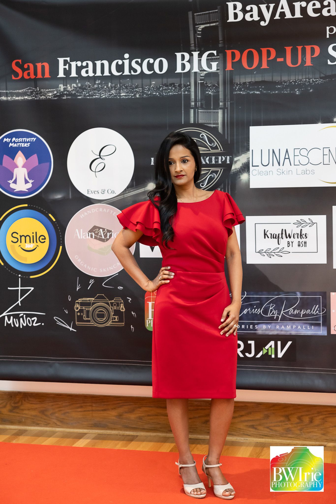 Woman poses in red cocktail dress with ruffle sleeves on red carpet at fashion event.