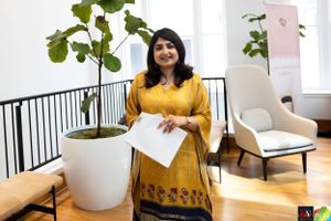 Woman in yellow ethnic dress stands next to large potted plant in modern interior space.