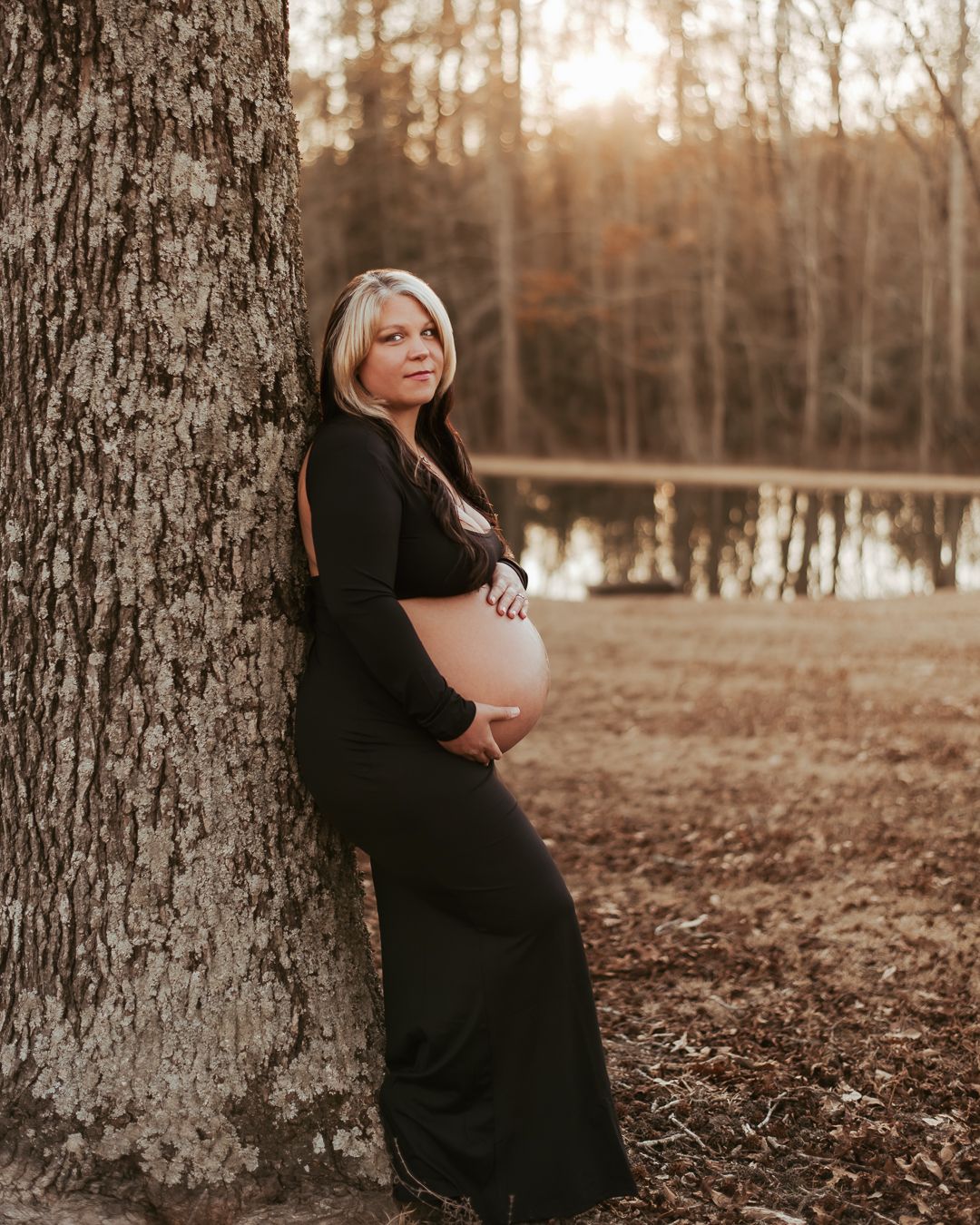 Maternity photo of woman in black dress leaning against large tree in autumn setting.
