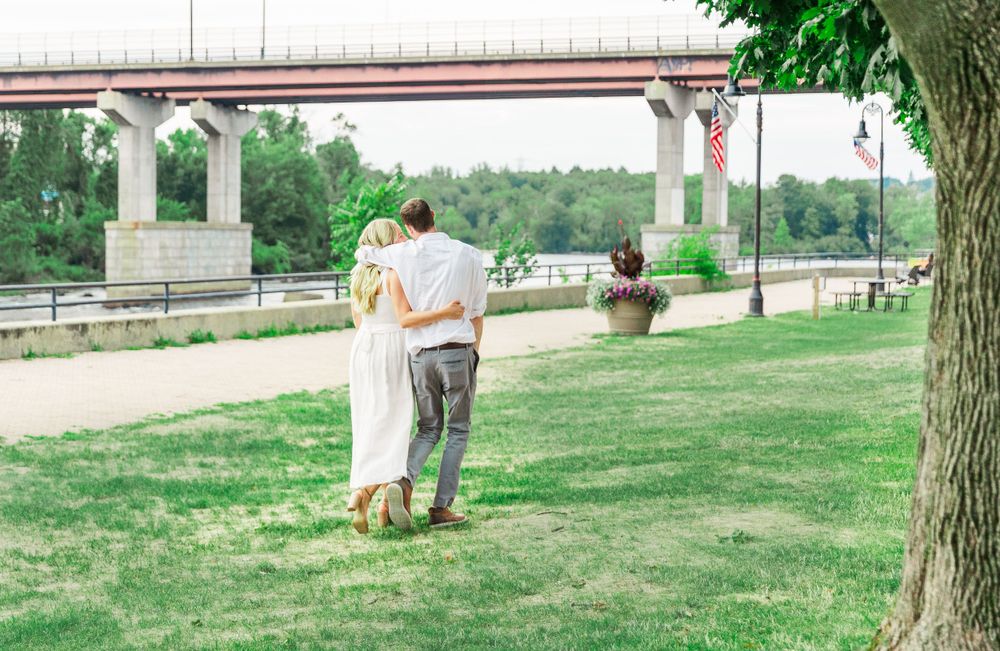 A couple embraces while standing on grass near a bridge.