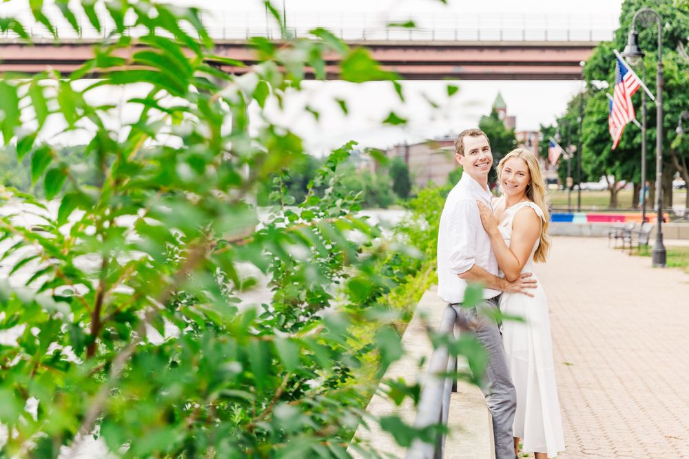 A couple poses together on a brick path with greenery and bridge in background.