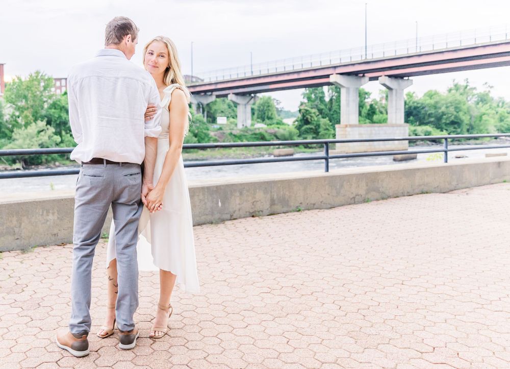 Couple in romantic pose standing on brick patio with bridge and greenery in background.
