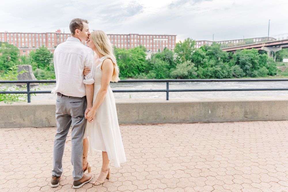 Romantic couple embracing on brick patio with industrial bridge in background.