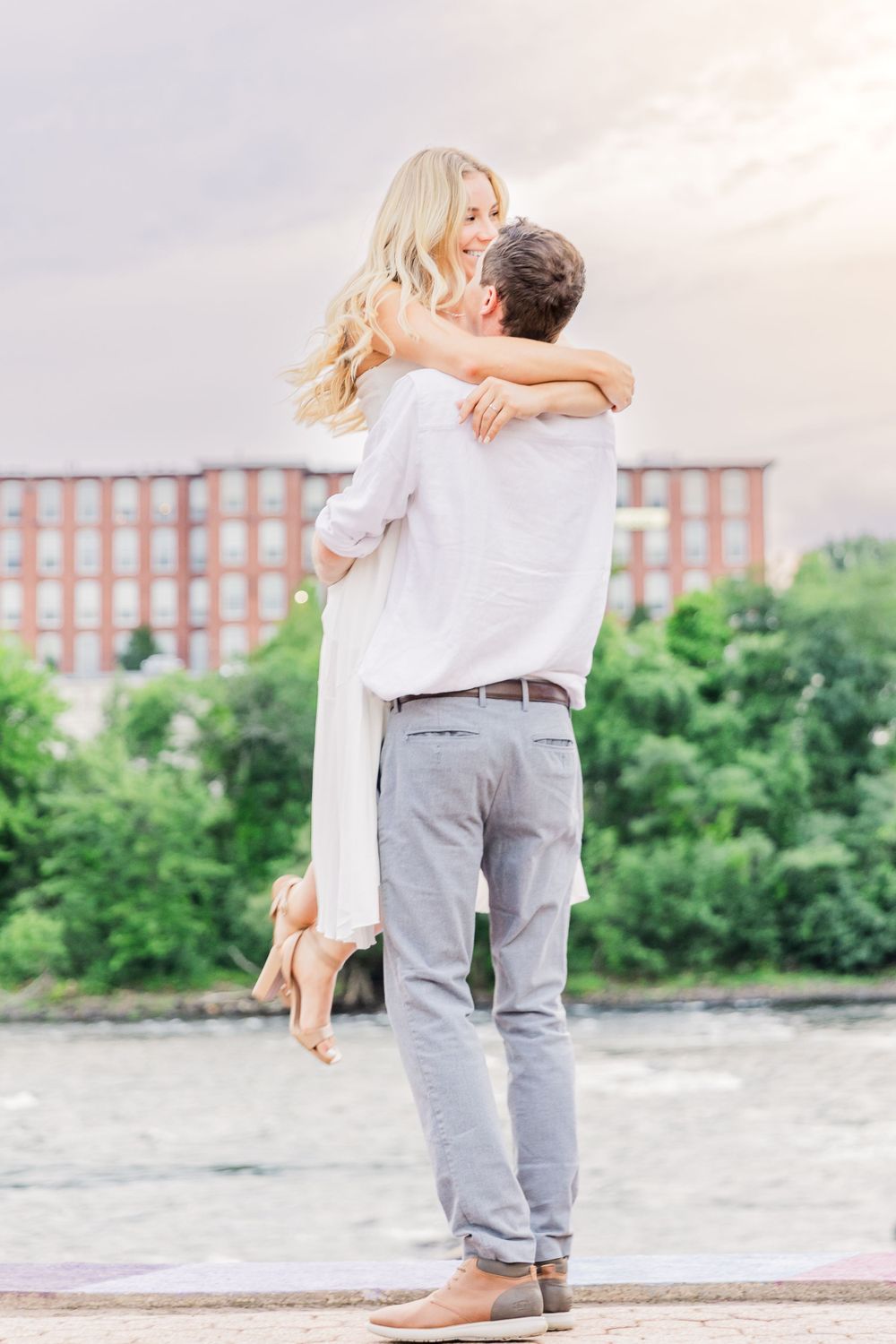 Man lifts woman in white dress during romantic photoshoot outdoors.