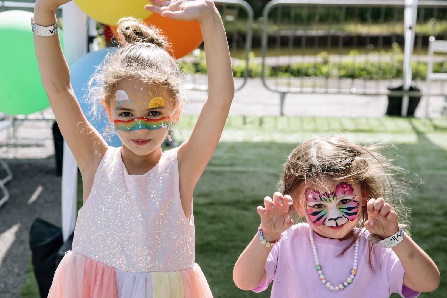 Two young children with face paint smiling and raising their arms at an outdoor event.