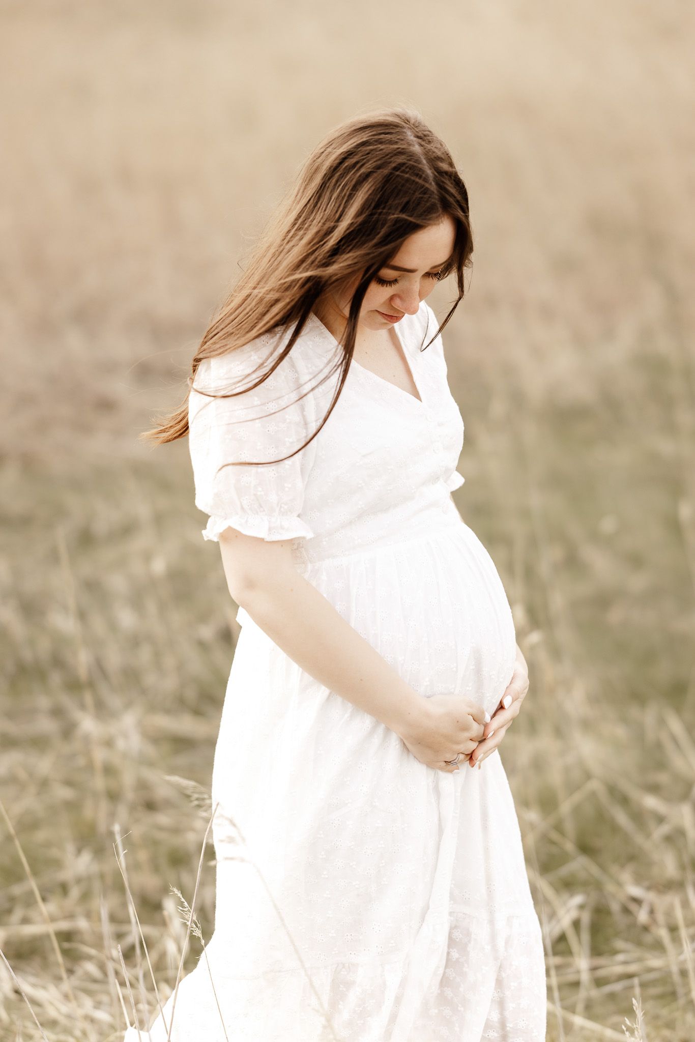 A woman in a flowing white dress cradles her baby bump in a serene field at golden hour.