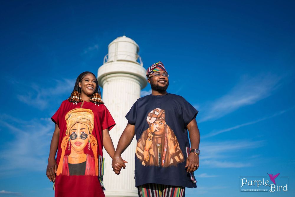 Couple in matching red and navy graphic tees holding hands and smiling in front of a white lighthouse under a bright blue sky.