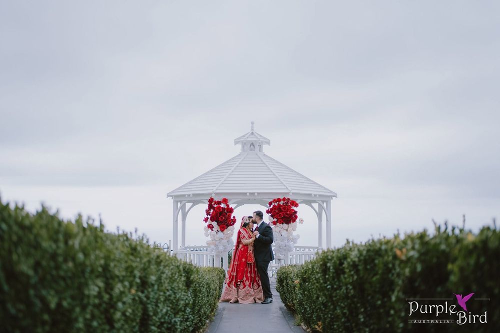 Couple in red and dark attire embrace beneath a white gazebo with red floral arrangements, framed by green hedges on a cloudy wedding day.