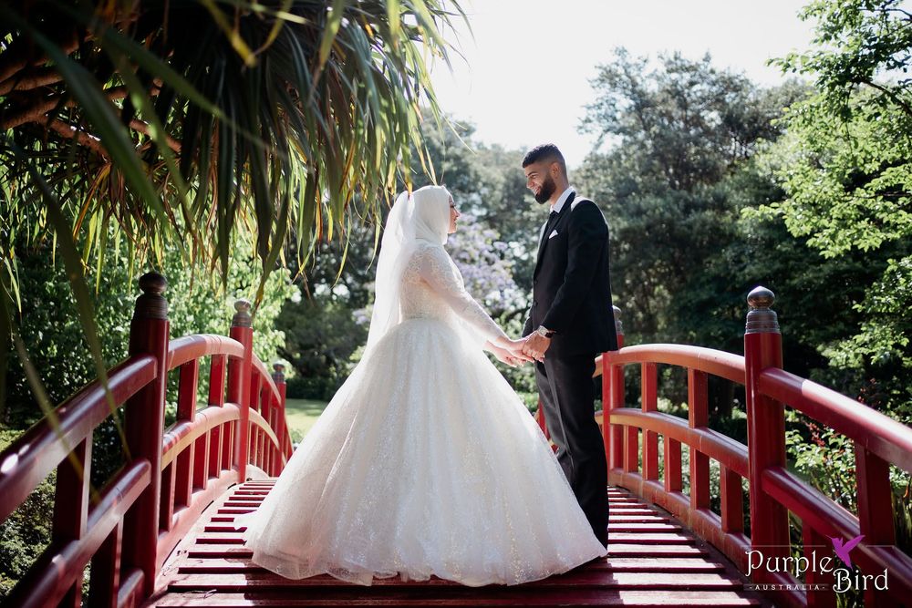 Bride in a white gown and veil holds hands with the groom in a dark suit on a red wooden bridge surrounded by lush green garden trees.