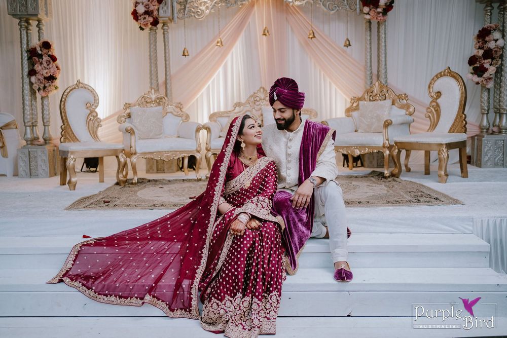 Indian bride in a maroon embroidered lehenga and groom in a matching purple turban sit together on white stairs in an elegant wedding venue.