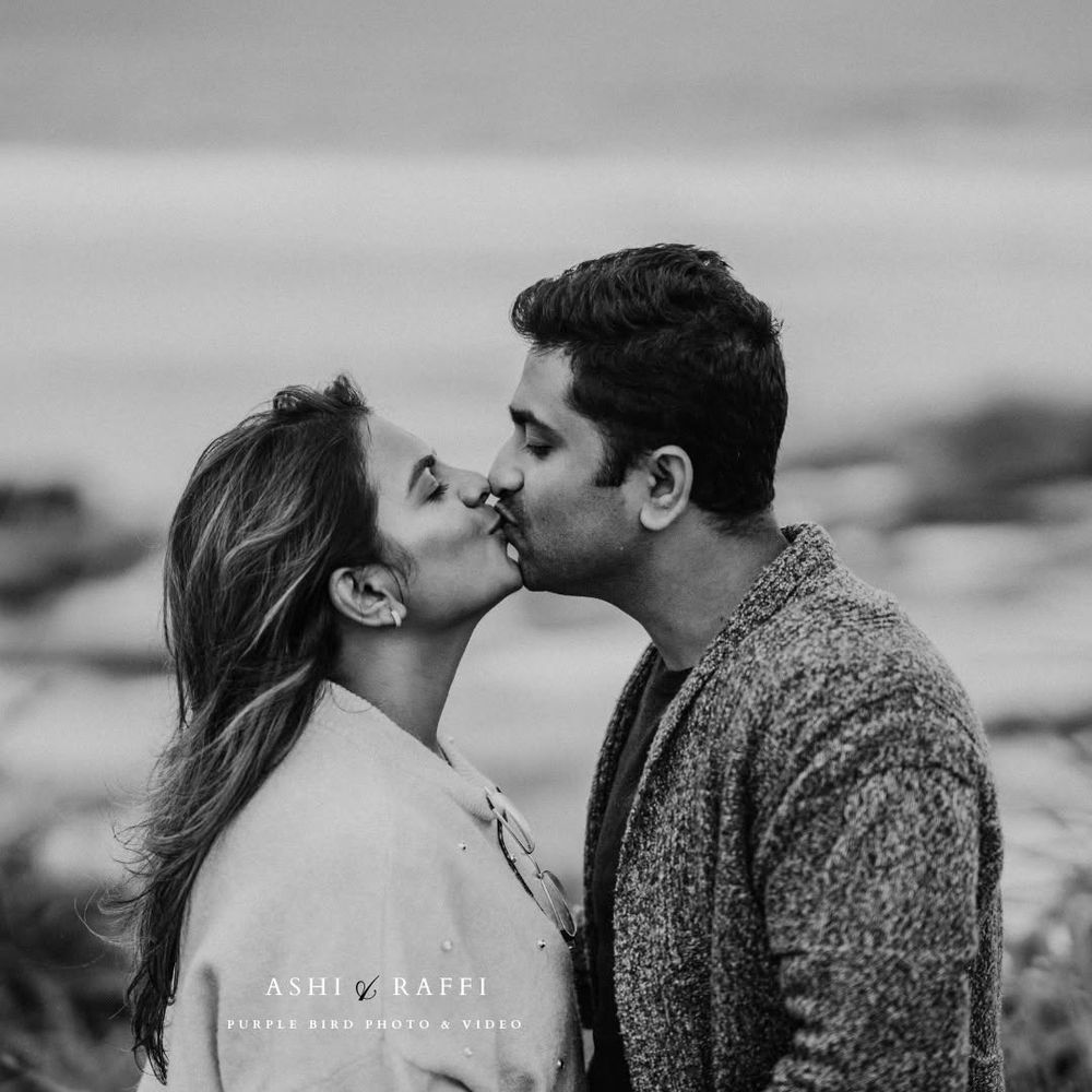 Black and white engagement photo of a couple sharing a kiss by the ocean, the woman in a light sweater and the man in a knit cardigan.