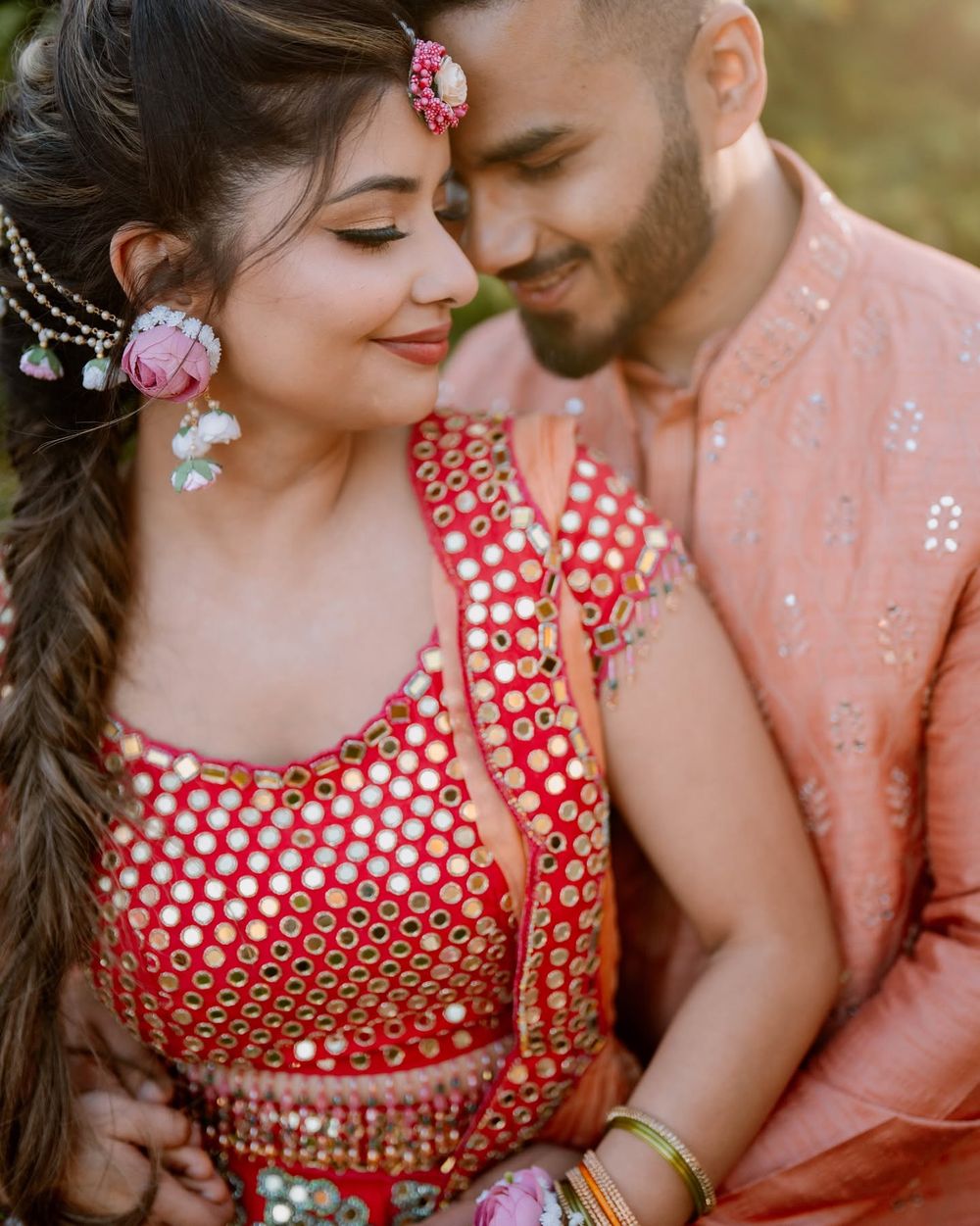 Smiling couple embracing in traditional Indian attire, she in a red mirror-work outfit with floral jewelry, he in a peach embroidered kurta.