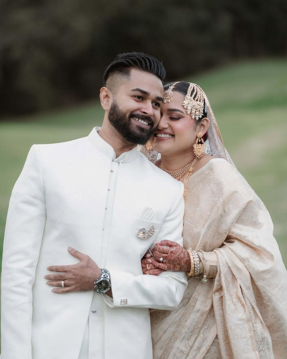 Smiling South Asian couple embracing outdoors, the groom in a white sherwani and the bride in a gold saree with henna and jewelry.
