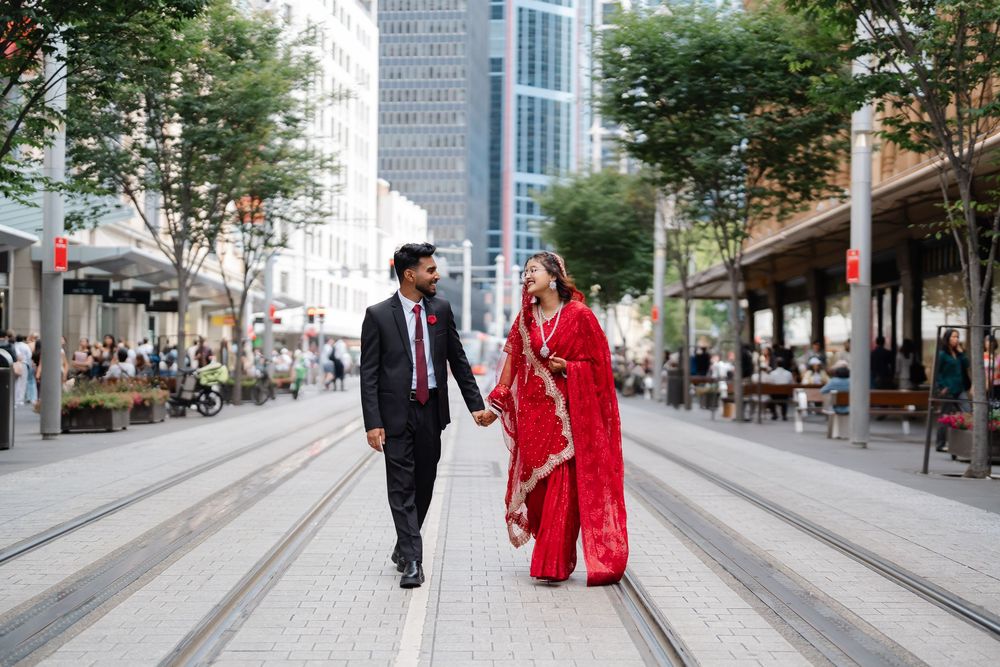 Smiling couple holding hands, the man in a dark suit and the woman in a red embroidered sari, walking along a tram-lined city street.
