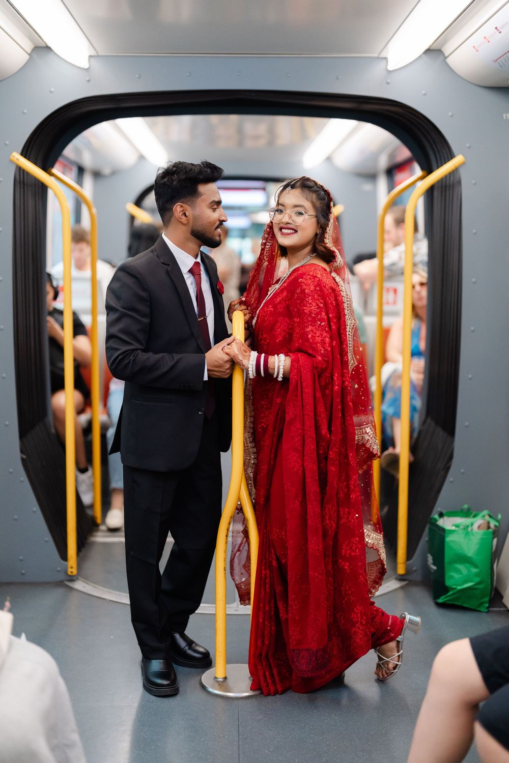 Bride in a red embroidered saree and groom in a dark suit hold hands beside a yellow pole inside a subway train carriage with passengers.