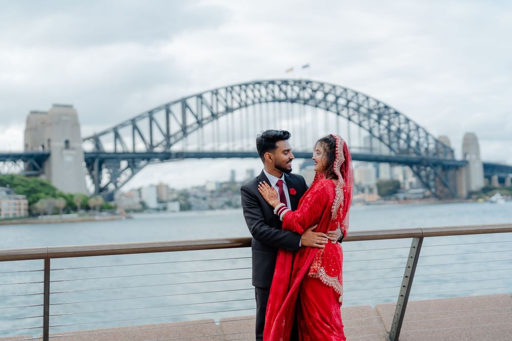 Couple in an embrace on a waterfront promenade, the bride in a red sari, with a large steel arch bridge and cloudy sky behind them.