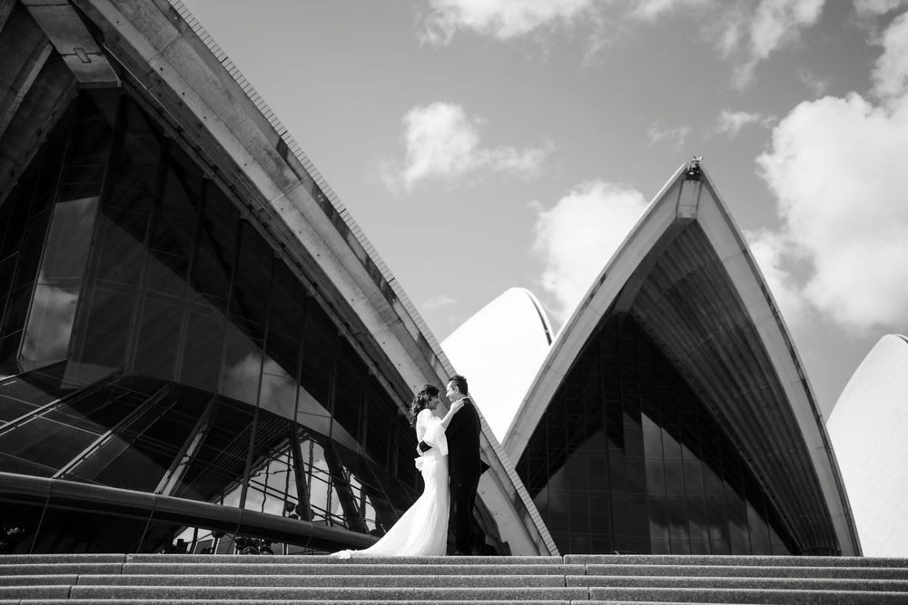 Black and white photo of a bride and groom embracing on the steps in front of the Sydney Opera House sail-shaped shells.