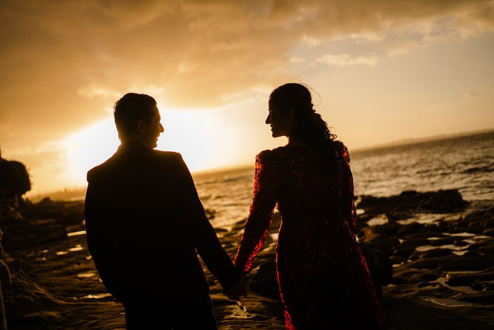 Silhouetted couple holding hands on a rocky shore, facing the ocean as the golden sunset glows across the sky.