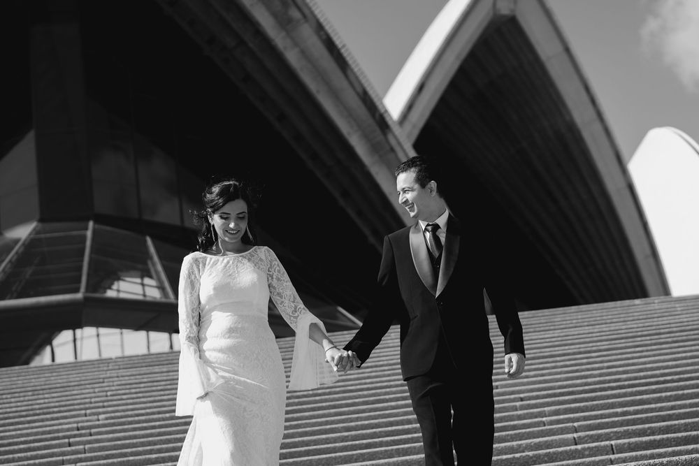 Black and white photo of a bride in a long-sleeved lace gown holding hands with a groom in a suit on steps near the Sydney Opera House.