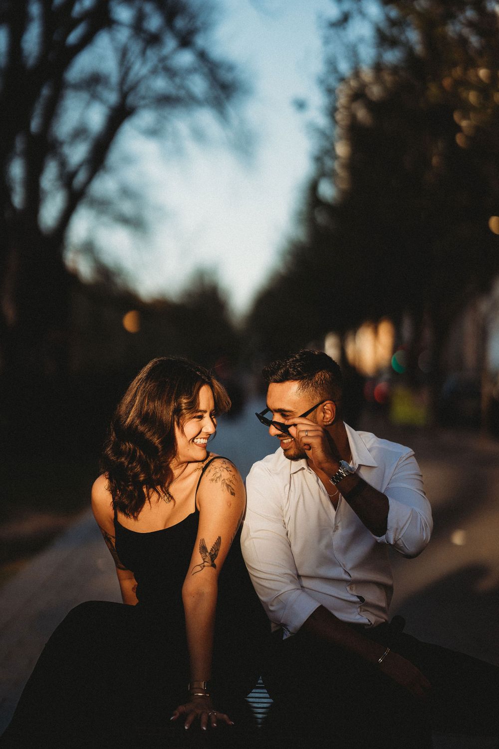 Couple sitting together at dusk on an empty street with trees silhouetted against the sky.