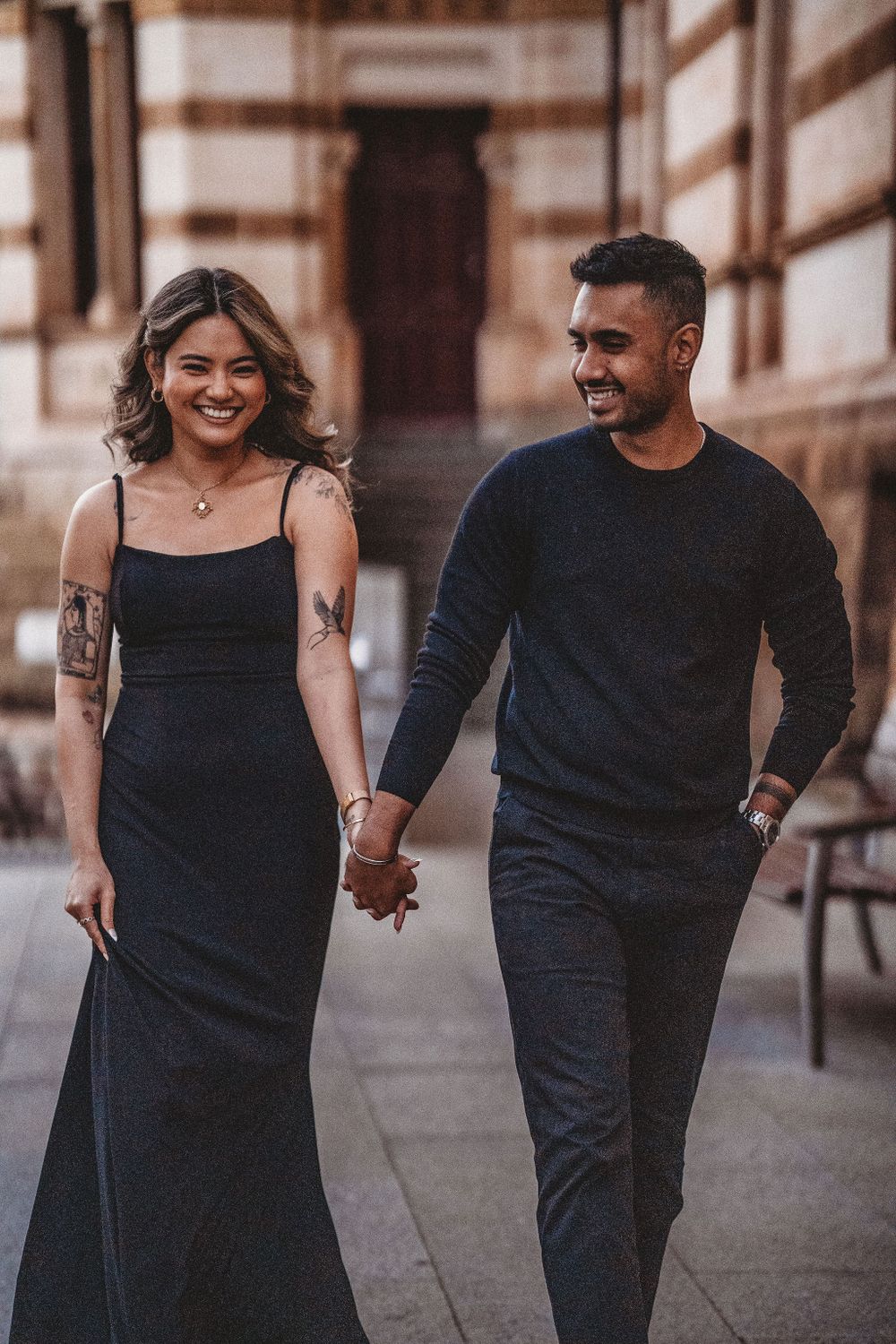 Couple in matching black outfits walking together along a historic building.