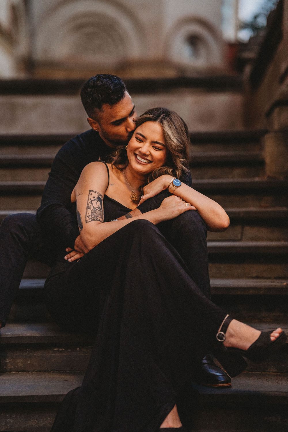Intimate couple in black formal wear embracing on stone steps with vintage architecture backdrop.