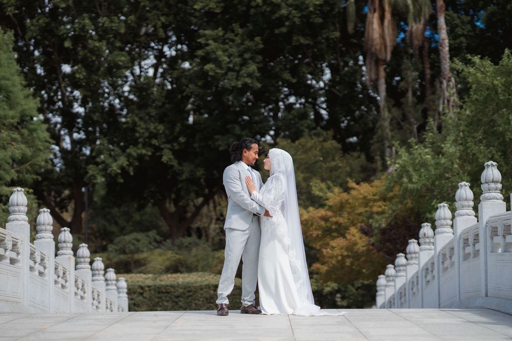 Bride in a white gown and veil embraces her groom in a gray suit on a white stone bridge with balustrades, surrounded by lush green trees.