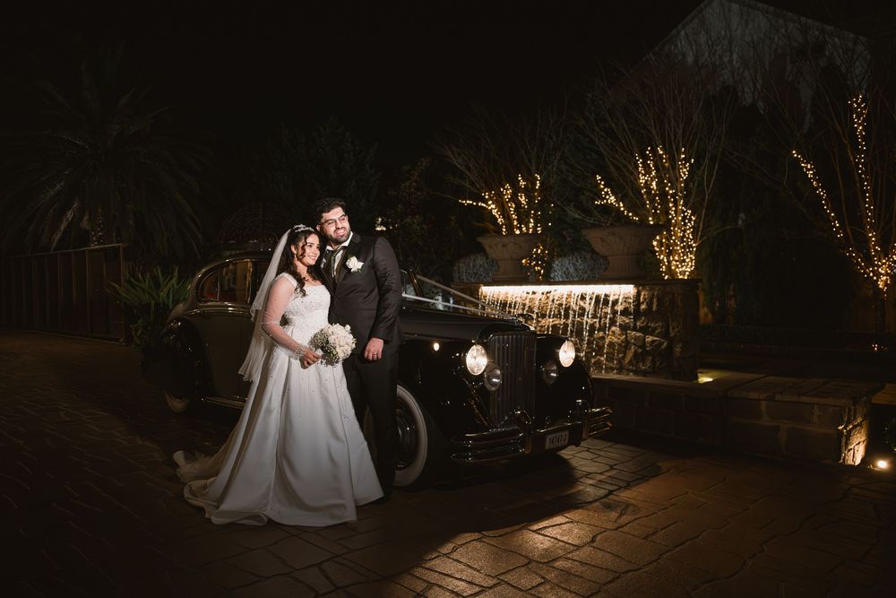 Bride in a white gown and groom in a dark suit pose beside a vintage black car at night, with glowing string lights behind them.
