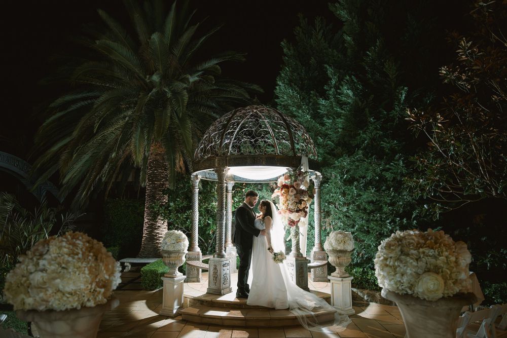 Bride and groom embrace inside a lit ornate gazebo at night, surrounded by palm trees and white floral urns at an elegant garden wedding.
