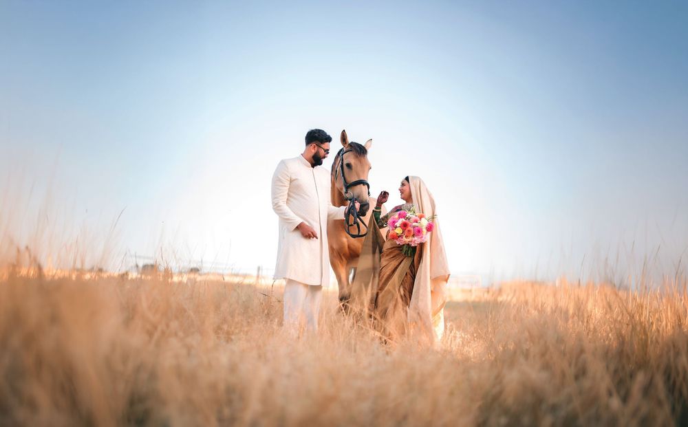 Couple in white traditional outfits stand beside a brown horse in a golden grass field under a soft blue sky at sunset.