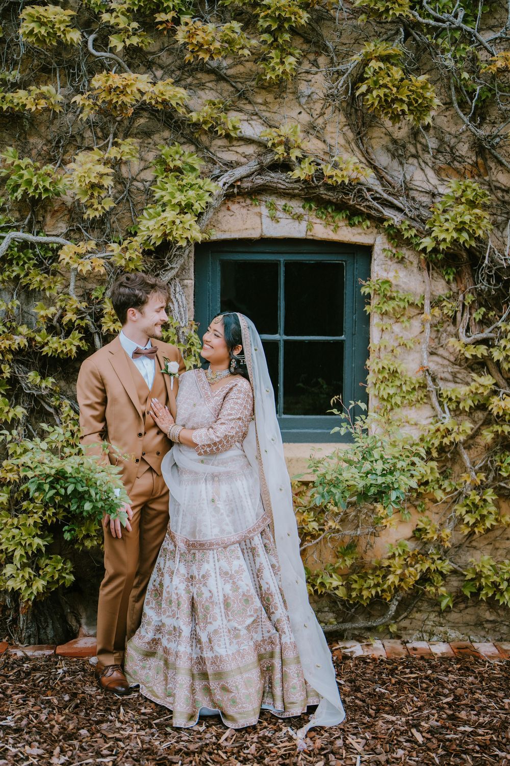 Bride in an ivory embroidered lehenga and veil embraces a groom in a tan suit beside an ivy-covered stone wall and window.