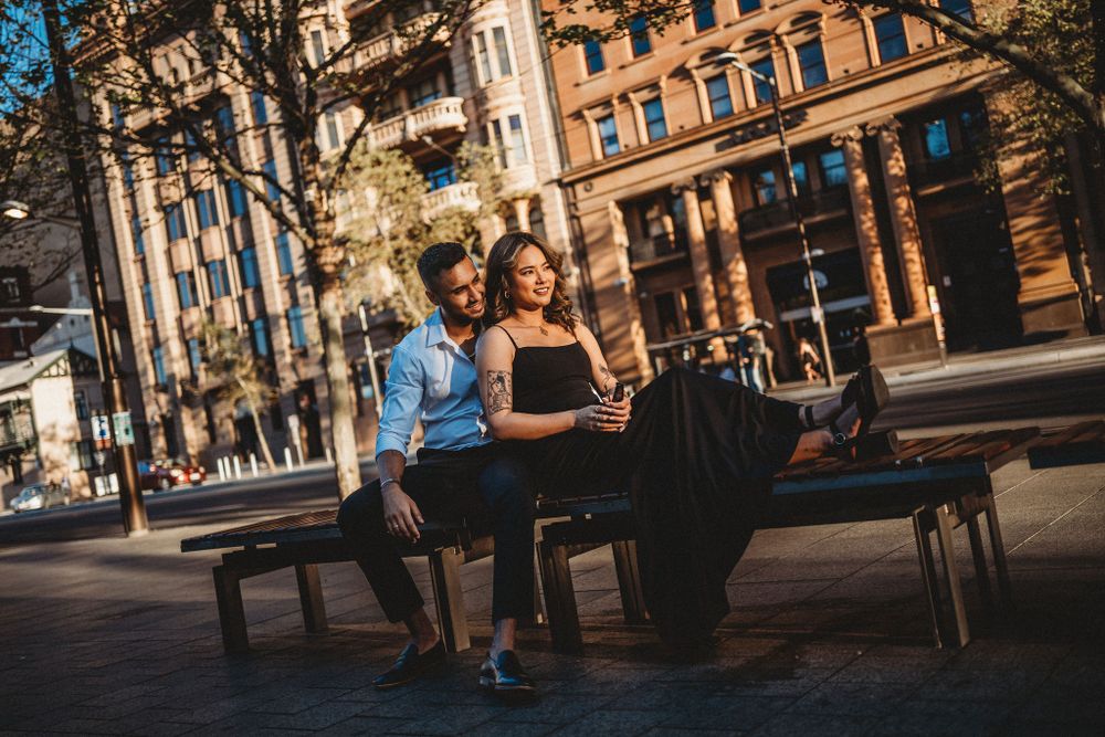 A romantic couple sits together on a bench outside a historic building on a sunny day.