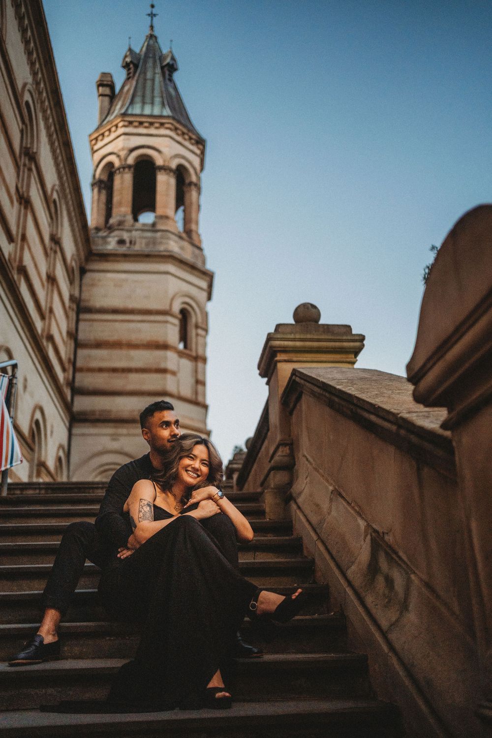 A couple sits on stone steps with a beautiful church spire towering in the background.