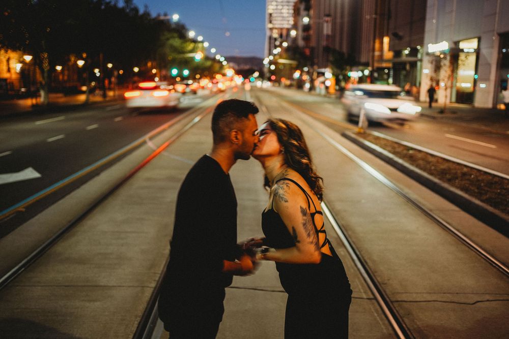 A romantic nighttime kiss on train tracks with city lights glowing in the background.