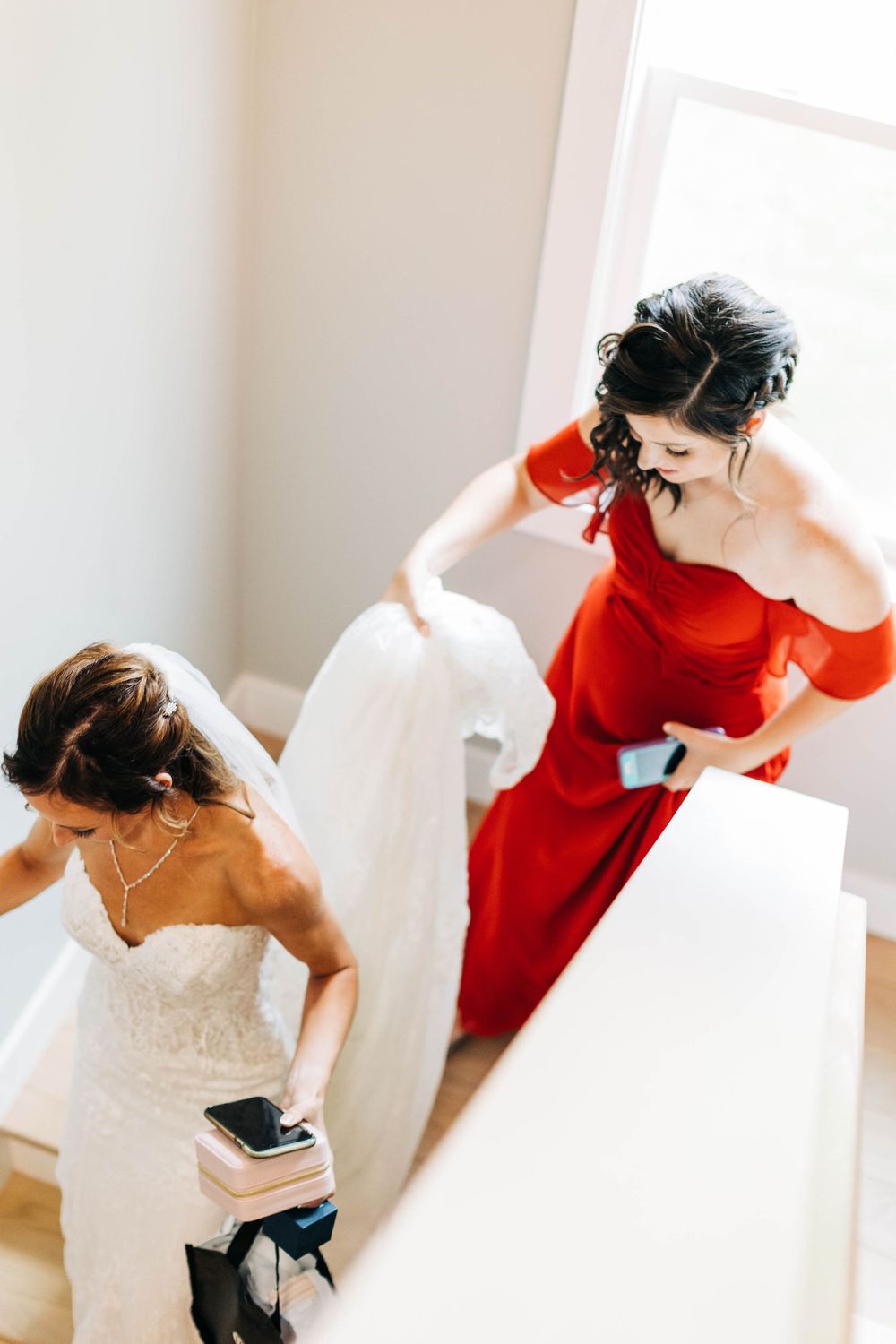 Woman in red dress helps adjust wedding gown in bright indoor setting.