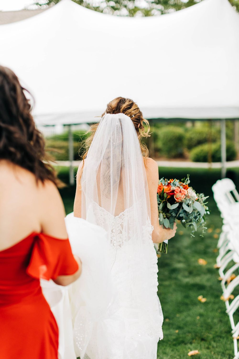 Outdoor wedding scene with flowing white veil and red bridesmaid dress against garden backdrop.