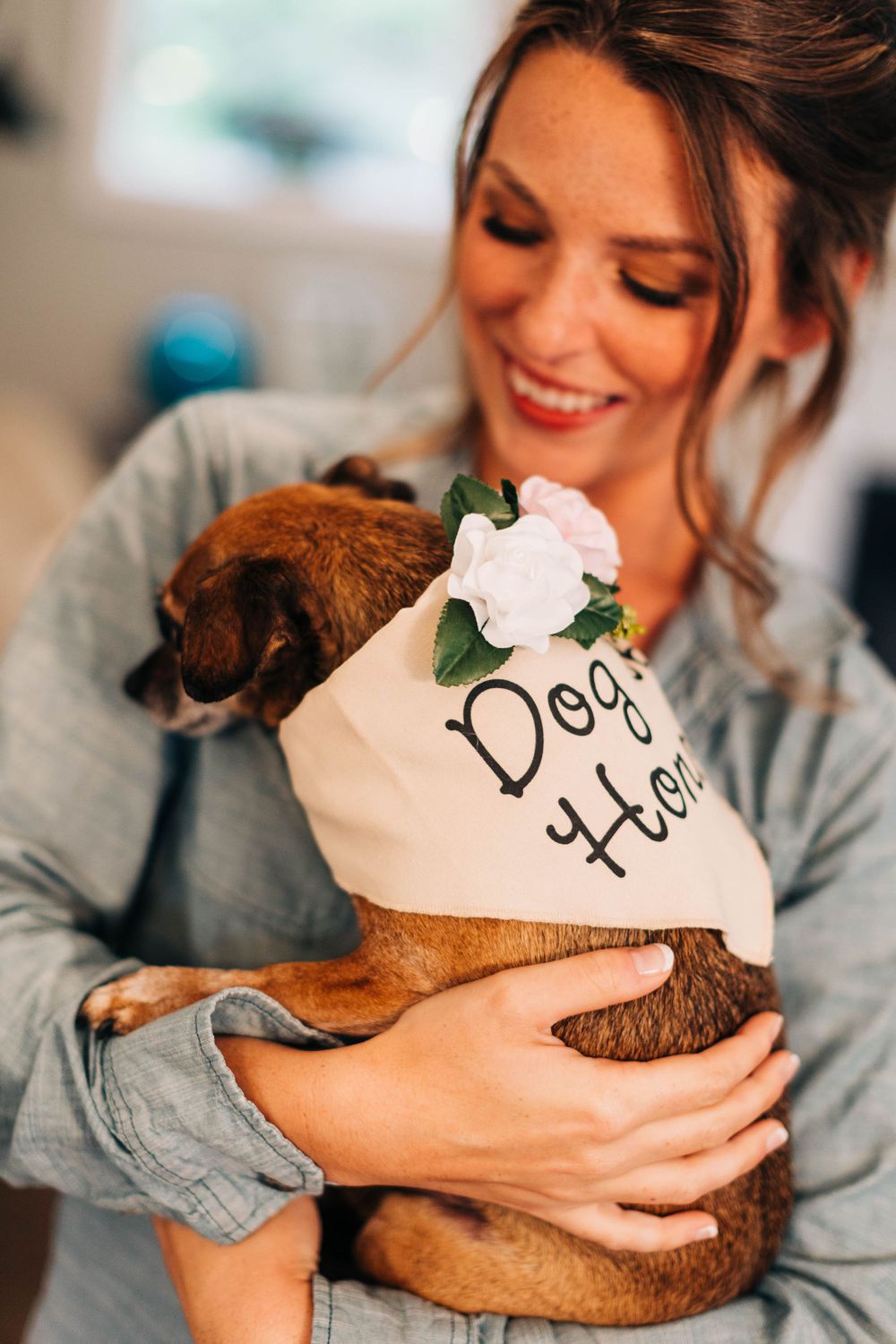 Sweet brown dog wearing a decorative bandana is held lovingly in a casual indoor setting.