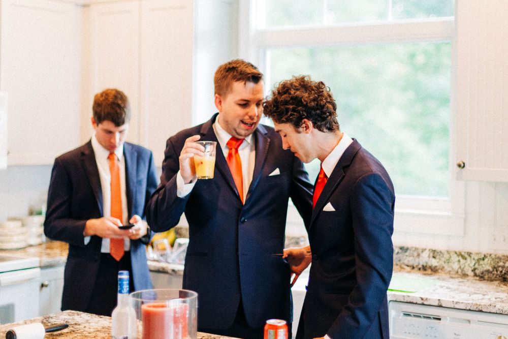 Groomsmen in navy suits and orange ties share drinks and laughs at a wedding reception.