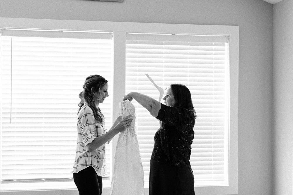 Two people examine a white wedding dress by a window in black and white photography.
