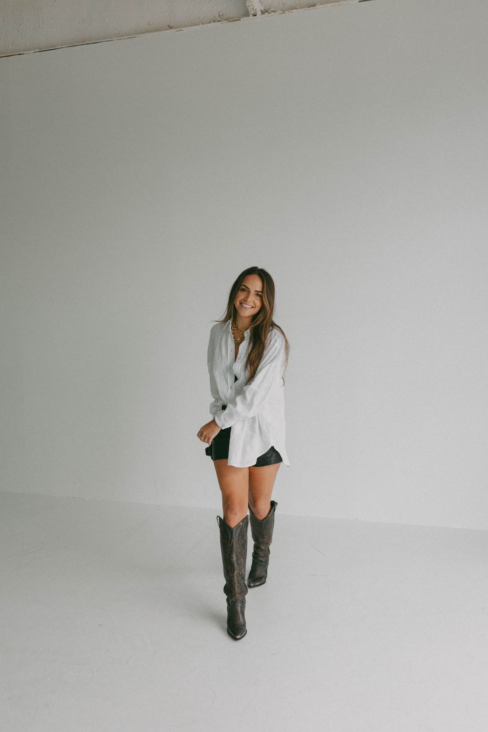 Woman in casual chic outfit with knee-high boots poses against white studio backdrop.