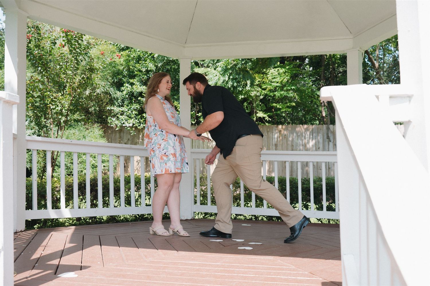 A couple shares a playful moment while dancing together on a white gazebo deck surrounded by greenery.