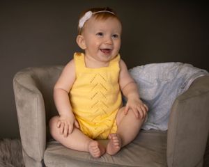 A baby girl sits on a small couch smiling