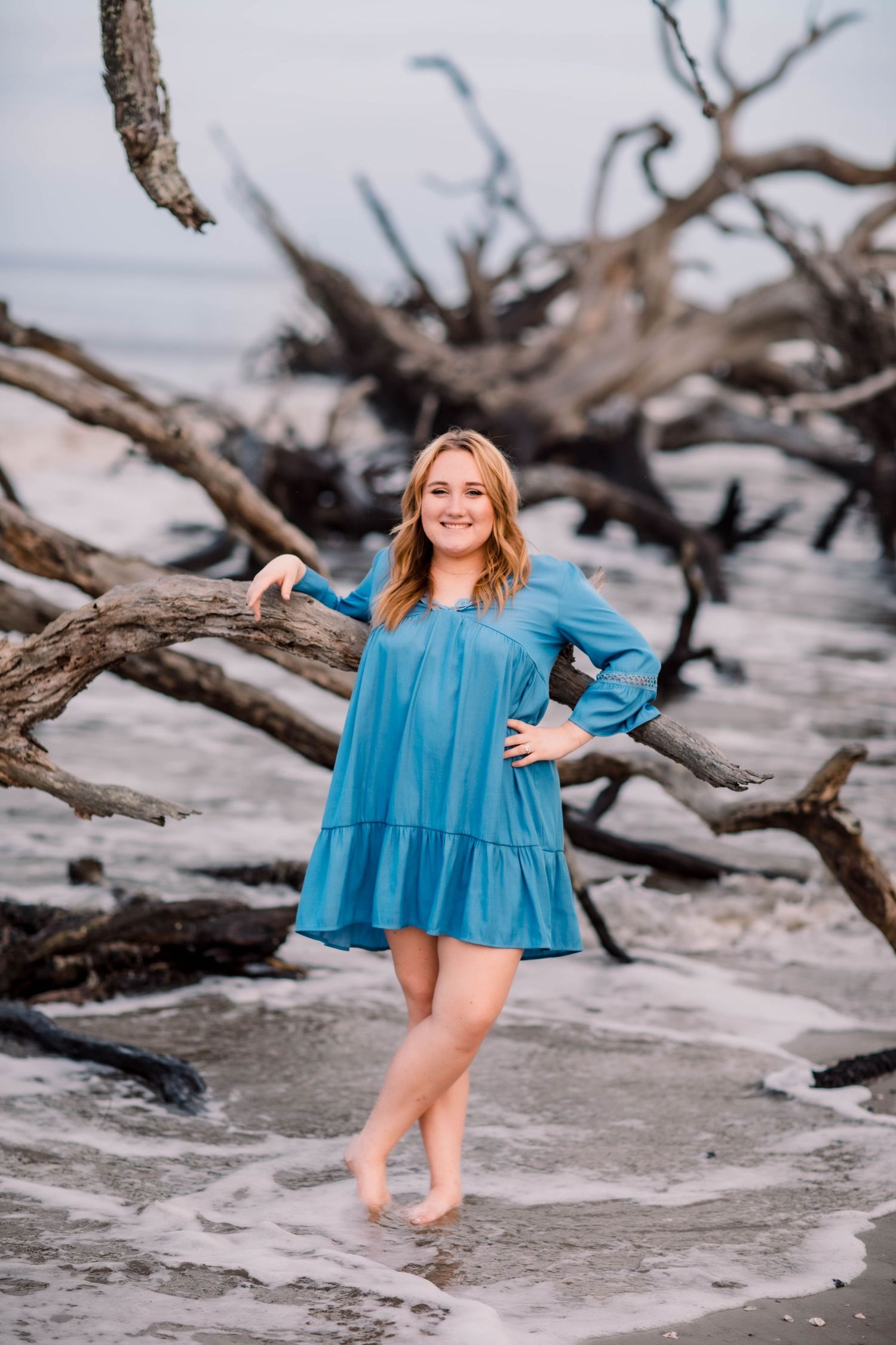 Woman in flowing blue dress posing on driftwood beach.