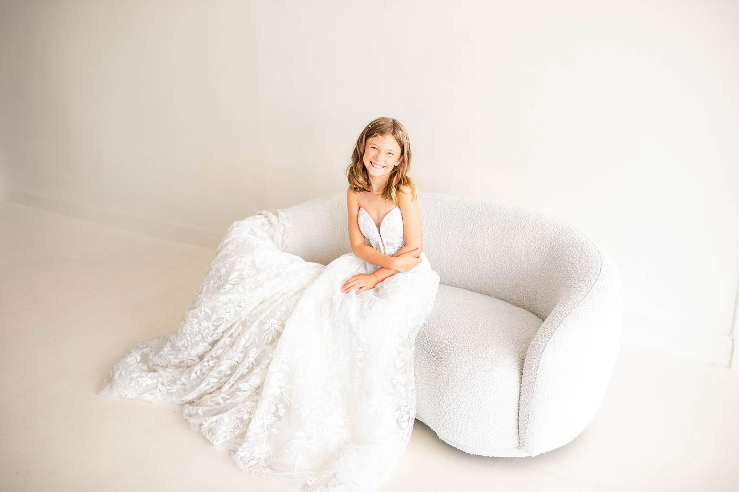 A bridal gown with delicate lace details and a flowing train is displayed in an elegant white studio setting.