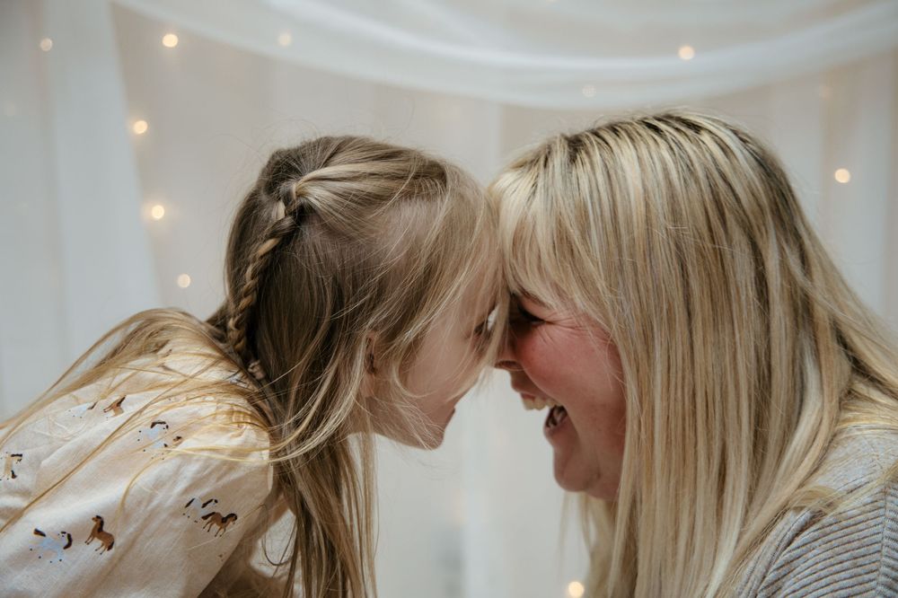 Laughing blonde mother and daughter touching foreheads playfully under fairy lights in a white tent.