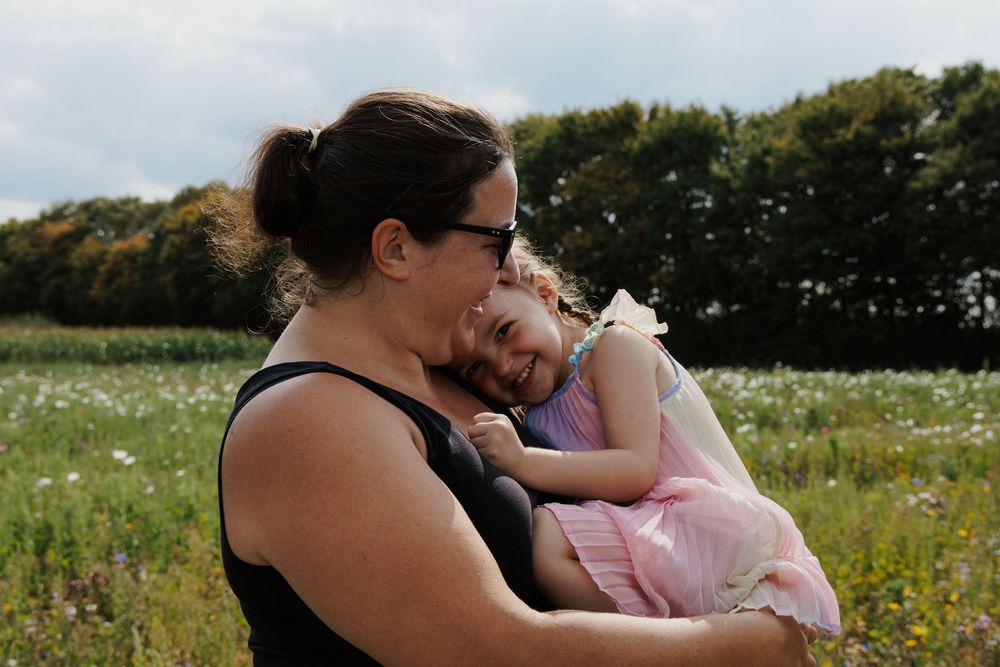 Mother lovingly embracing her smiling daughter in a pink dress in a sunny wildflower field.