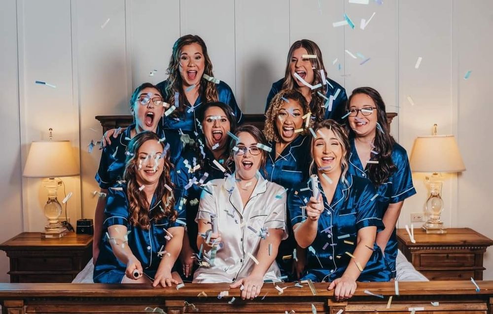 Bridesmaids in navy satin pajamas celebrate with confetti poppers alongside bride in white during a fun bridal party getting-ready moment.