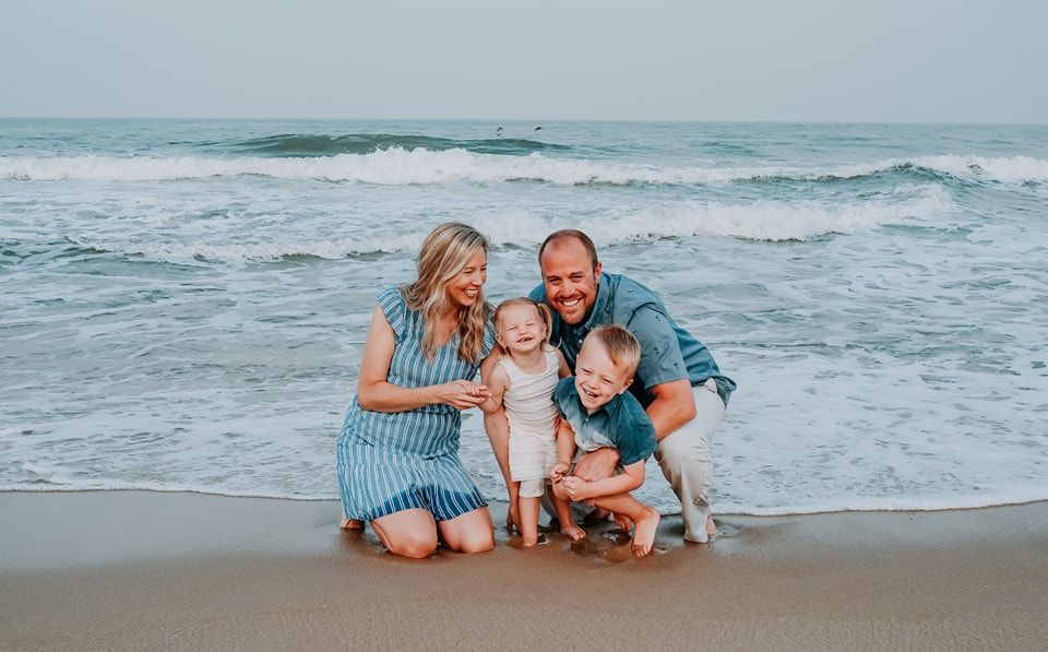 Happy family of four smiling together on a sandy beach with ocean waves in the background.