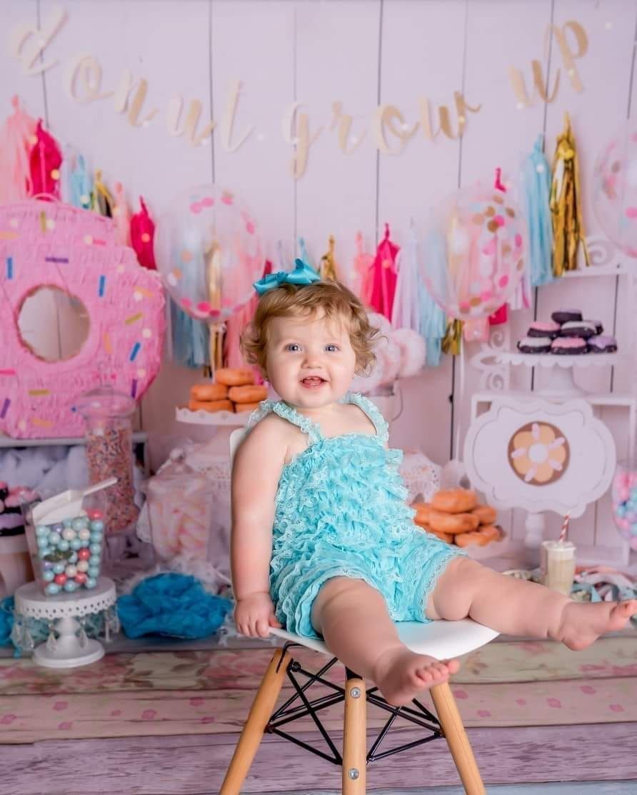 Adorable baby girl in teal romper sits on white chair at donut-themed first birthday party celebration.