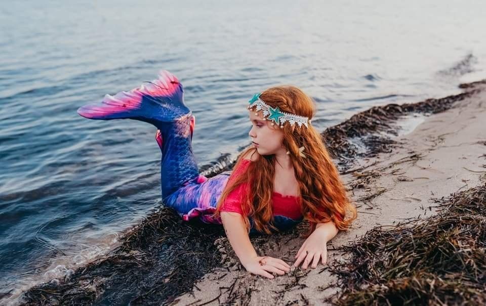A redhead woman in a mermaid costume with colorful tail and floral crown poses on a rocky beach shoreline.